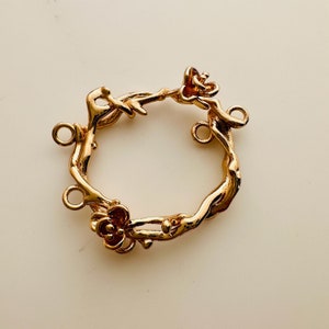 May include: A gold-coloured metal wreath-shaped jewellery finding. The wreath is designed to resemble intertwined branches with small flower accents. It has four loops for attaching to other jewellery components.