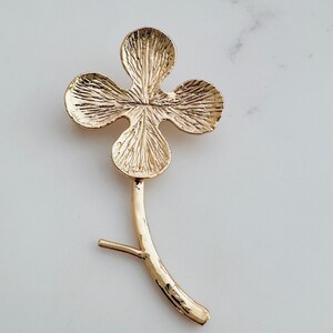 May include: A gold-tone clover leaf brooch with a stem. The clover has four leaves with a textured surface.