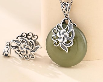 Può includere: Una collana con pendente in argento con una grande pietra ovale verde oliva. Il pendente è adornato da un motivo floreale argentato. È visibile anche un anello in argento abbinato con un motivo floreale simile.