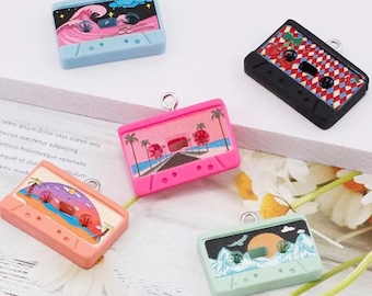 May include: Five decorative cassette tape charms in various colours. One is light blue with a wave design, another is pink with a beach scene, and a third is orange with a desert scene. The other two are black and mint green.
