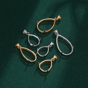 May include: A selection of teardrop-shaped pendant settings in gold and silver. The settings have a loop at the top for a chain and small prongs to hold a gemstone. The pendants are displayed on a dark green background.