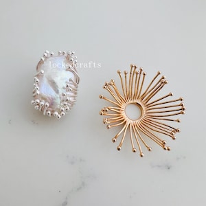 Könnte beinhalten: Zwei Schmuckstücke, eines ist ein silberner Ring mit einer großen weißen Perle, das andere ist ein goldener Ring mit einem kreisförmigen Design aus dünnen Drähten, die nach außen strahlen.