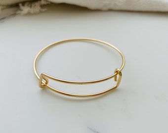 May include: A gold-colored adjustable bangle bracelet. The bracelet features a circular design with a rectangular adjustable section. The metal has a shiny finish. The bracelet is shown on a white marble surface.