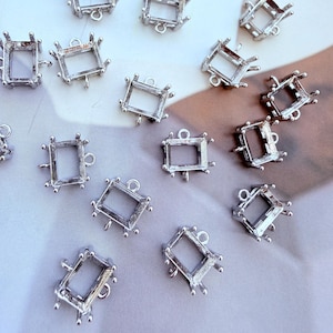 May include: A collection of silver-toned metal rectangular jewellery settings, each featuring four prongs at the corners and a small circular loop at the top for attachment. These open-back frames are designed to hold gemstones or cabochons.