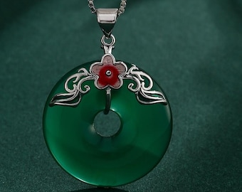 Può includere: Collana con pendente in giada verde con catena d'argento. Il pendente presenta una pietra di giada circolare con un foro centrale, ornata da un fiore rosso e dettagli in filigrana argentata. La collana è su sfondo verde scuro.