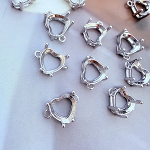 May include: A collection of silver-toned metal heart-shaped jewellery settings. Each piece features a hollow centre, raised prongs for securing a gemstone, and a small circular loop at the top for attachment, designed for crafting necklaces or earrings.