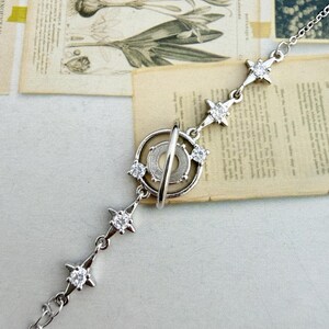 May include: A silver-toned bracelet featuring a celestial design. The bracelet has a central circular element with a bar, and is adorned with star-shaped accents set with clear gemstones. The chain is delicate and adjustable.