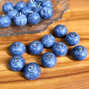 May include: Blue blueberry-shaped beads with silver metal loops, scattered on a wooden surface and in a clear dish. The beads are a deep blue colour and have a textured surface. The beads are approximately 2 cm in diameter.