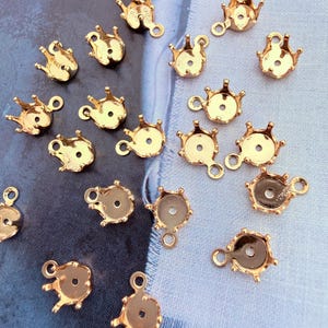 May include: A collection of small, gold-coloured metal jewellery settings, each featuring a crown-like prong design with a circular base and a small loop at the top for attachment. These components are arranged in a scattered pattern.