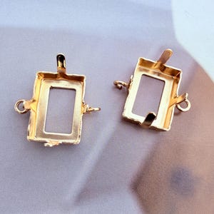 May include: Two gold-coloured rectangular jewellery settings with open centres. Each setting features four small prongs extending from the edges and two small circular loops on opposite sides for attachment to chains or other jewellery components.