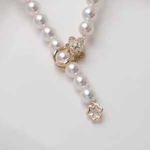 May include: A white pearl necklace with a gold flower clasp and a gold flower pendant. The necklace is on a white surface.
