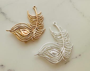 May include: Two peacock feather charms, one gold and one silver, with a detailed design and a loop at the top for attaching to jewellery or crafts.