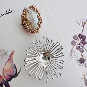 Peut inclure: Deux bijoux, l'un est une bague dorée avec une grosse perle blanche, l'autre est une broche en forme de fleur argentée avec un centre rond.