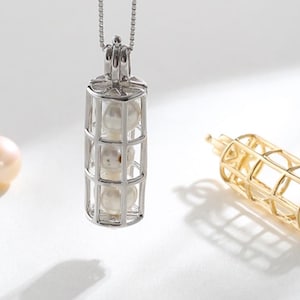 May include: A silver pendant necklace with a cage design, holding three pearls. A loose pearl and a gold-colored cage pendant are also present. The jewelry is set against a white background, highlighting the elegant design.