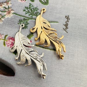 May include: Two metal feather charms, one silver and one gold, sit on a grey floral patterned surface. The charms have detailed vein patterns and a loop at the top for hanging. The gold charm is shiny, while the silver charm has a matte finish.