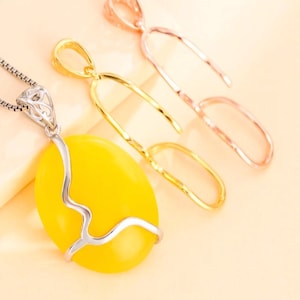 Op de afbeelding: Een zilveren ketting met een grote, ovale, gele hanger. De hanger is geaccentueerd met een zilveren, golvend ontwerp. Twee gouden en roségouden hangerblanks zijn op de achtergrond.