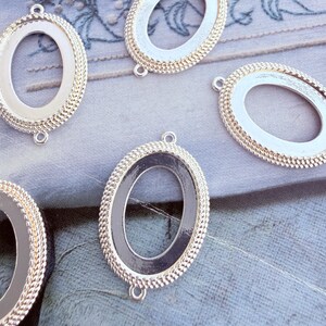 Peut inclure: Quatre connecteurs de bijoux en métal de forme ovale avec une finition argentée. Chaque pièce présente une bordure texturée en forme de corde entourant un centre lisse et encastré. De petites boucles sont situées en haut et en bas.