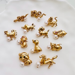 May include: Twelve gold animal charms with red accents and white pearl accents. The charms are shaped like a rooster, snake, tiger, pig, horse, dog, monkey, ram, rabbit, mouse, dragon, and a fish.