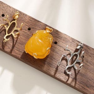 May include: Three pendants on a wooden surface. One is a gold-colored abstract design, another is a silver-colored abstract design, and the third is a yellow-orange stone pendant with gold-colored accents. The pendants are for jewelry.