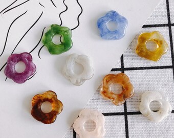 May include: Assortment of eight flower-shaped resin charms in various colors, including purple, green, blue, brown, white, and pink. Each charm has a hole in the center.