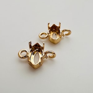 May include: Two gold-toned metal oval-shaped settings with loops for attaching to jewelry. The settings are designed to hold gemstones or other embellishments.