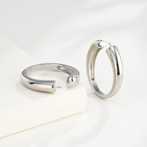 May include: Two silver-colored hoop earrings. One earring is open, displaying the clasp. The other earring is closed. The earrings have a smooth, rounded design and are set against a white background.