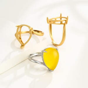 May include: Three rings are displayed against a white background. Two are gold-colored, with open settings. The third ring has a silver band and a large, yellow, teardrop-shaped stone.
