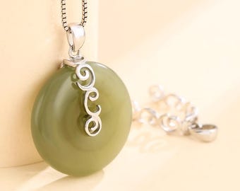 Può includere: Collana con pendente in giada verde oliva e catena d'argento. La pietra di giada ovale è completata da un'ansa decorativa in argento. La collana è presentata su uno sfondo beige chiaro.