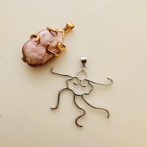 May include: Two pendants on a cream surface. One has a large, lavender-pink baroque pearl in gold-coloured metal. The other is a silver-coloured, abstract pendant with a dancer-like design.
