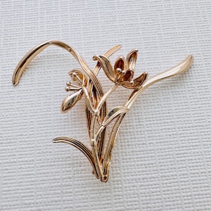May include: A gold-toned metal brooch in the shape of a stylized flower cluster. The brooch features delicate, intricate details and a three-dimensional design.