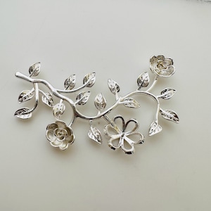 May include: A silver floral branch with leaves and three flowers. The design has a detailed, openwork style, with a polished finish. The leaves and flowers are intricately shaped, creating a delicate and elegant aesthetic.