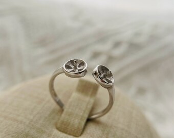 May include: A silver ring with two round, open-backed settings. The ring is designed to hold two small cabochons.