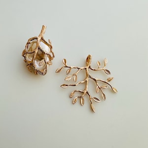 May include: Two gold-toned metal tree branch charms with intricate leaf details. The charm on the left is a closed tree branch with leaves, while the charm on the right is an open tree branch with leaves.