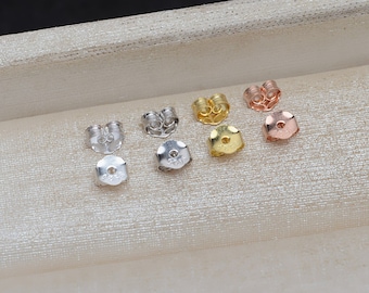 May include: A collection of earring backs in various metallic finishes, including silver, gold, and rose gold. The earring backs are displayed on a light beige surface, showcasing their different shapes and styles.