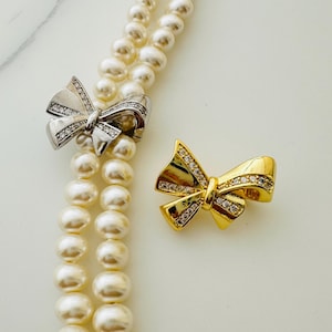 May include: Two pearl necklaces with a silver and gold bow pendant. Both bows are adorned with clear rhinestones.