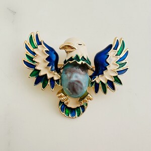May include: A gold-tone brooch featuring a stylized eagle with outstretched wings. The eagle is decorated with blue, green, and white enamel. The eagle's body is a light blue, green, and white marbled stone.