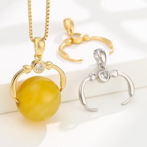 Puede incluir: Collar con colgante dorado con un orbe amarillo y dos colgantes adicionales. El colgante presenta un engaste dorado con una gema transparente y un diseño curvo que sujeta el orbe amarillo. Los otros colgantes son dorados y plateados.
