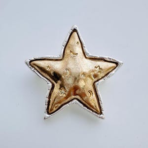 May include: A gold star-shaped brooch with a silver border. The star is embossed with a pattern of small stars and crescent moons.