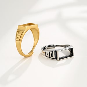 May include: Two rings, one gold-toned and one silver-toned, each featuring a rectangular bezel. The gold ring has a decorative geometric design on the band. The silver ring has a black geometric design on the band.