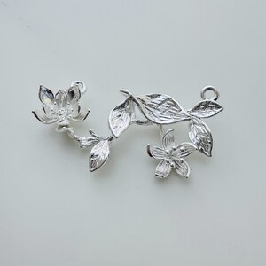 May include: Silver charm featuring a delicate floral design with two blossoms and leaves. The charm has a loop for attaching to a bracelet or necklace.