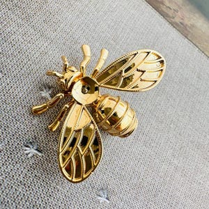 Könnte beinhalten: Eine goldfarbene Bienenbrosche mit detaillierten Flügeln, Körpersegmenten und Fühlern. Die Brosche besteht aus glänzendem Metall und ist auf einem grauen Stoffhintergrund platziert. Die Biene ist ein dekoratives Accessoire.