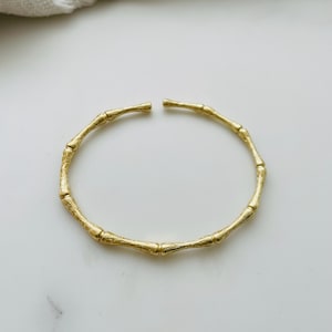 May include: A gold-toned bangle bracelet with a bamboo-inspired design. The bracelet is made of multiple connected pieces that resemble bamboo stalks.
