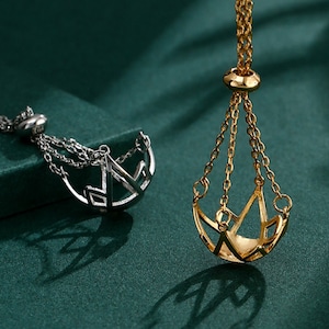 Könnte beinhalten: Zwei goldfarbene Halsketten mit geometrischem Design. Die Halsketten bestehen aus einer zarten Kette und haben einen Anhänger, der an eine Schaukel erinnert.