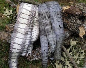May include: Three rolls of faux leather fabric with a snakeskin pattern. The rolls are in shades of white, gray, and dark gray. The fabric is draped over logs and grass.