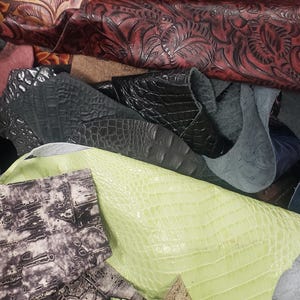 May include: Assortment of leather scraps in various colors and textures. Includes embossed floral patterns in shades of orange and red, black crocodile-embossed leather, and pale green alligator-embossed leather. Also includes a gray and white patterned leather.
