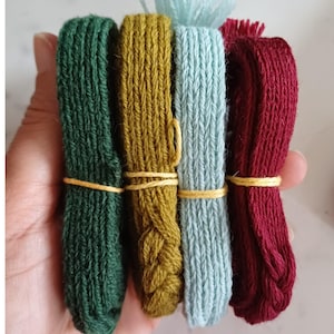 May include: Four bundles of colorful embroidery floss in shades of green, gold, light blue, and burgundy. Each bundle is secured with a thin yellow band. The floss is tightly wound, showcasing the texture and color variations.