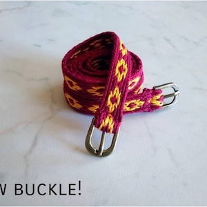 May include: A woven belt with a burgundy and yellow pattern and silver buckles. "NEW BUCKLE!" is written in black text below the belt.
