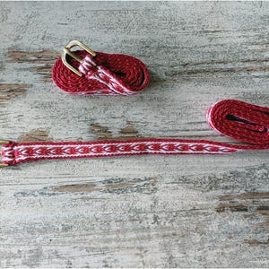 May include: A red and white woven belt with a brass buckle. The belt is coiled up and laid out flat on a wooden surface.