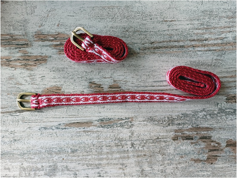 May include: A red and white woven belt with a brass buckle. The belt is coiled up and laid out flat on a wooden surface.