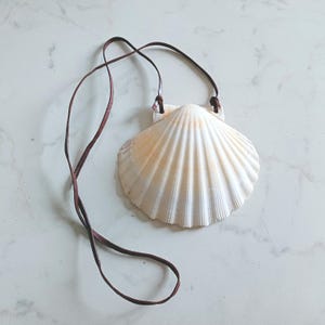 May include: A natural seashell pendant with a brown leather cord. The large, white shell has a ridged texture and is attached to the cord with small leather pieces. The pendant is a simple, beach-themed accessory.